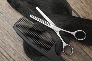 Black hair strand, comb and professional scissors on wooden surface, flat lay