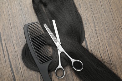 Photo of Black hair strand, comb and professional scissors on wooden surface, flat lay