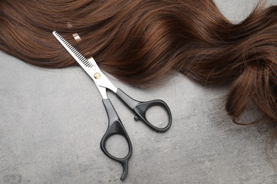 Photo of Brown hair strand and professional scissors on grey surface, flat lay