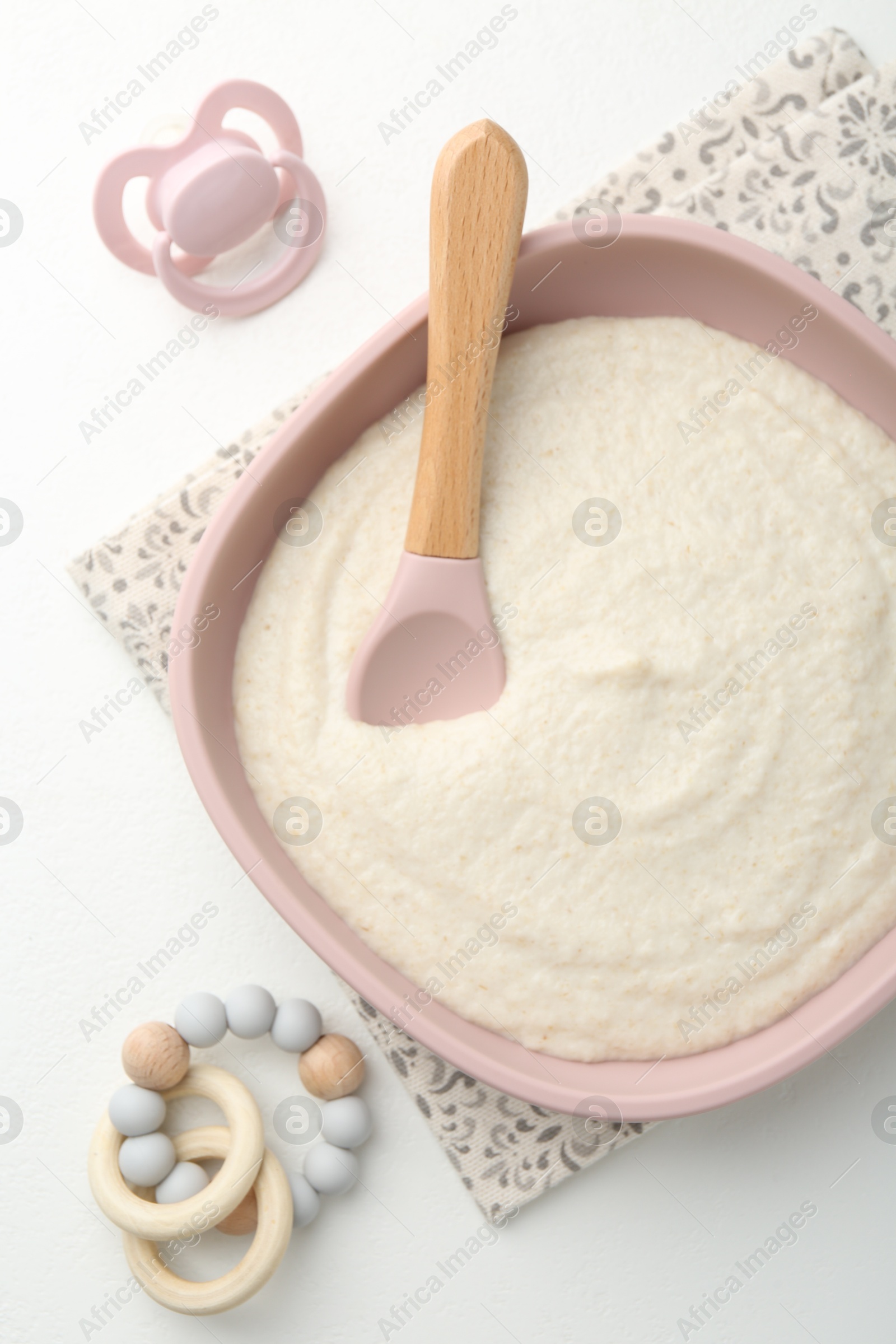 Photo of Delicious baby food in bowl with spoon, pacifier and teether on white background, flat lay