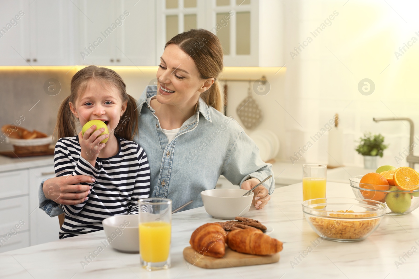 Image of Mother and her cute little daughter having breakfast at table in kitchen on sunny morning