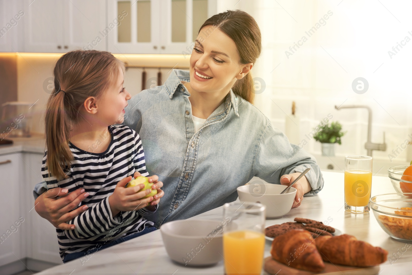 Image of Mother and her cute little daughter having breakfast at table in kitchen on sunny morning