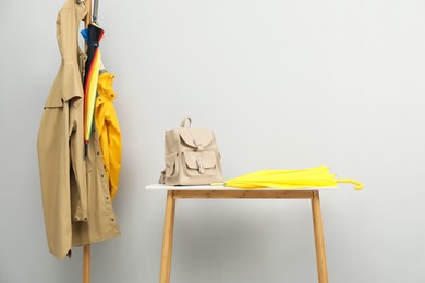 Photo of Yellow umbrella and backpack on white table near rack with coats against light grey background