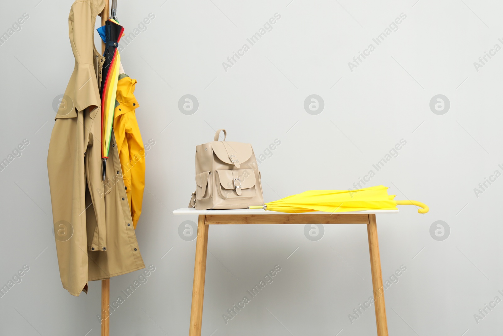 Photo of Yellow umbrella and backpack on storage bench near rack with coats against light grey background