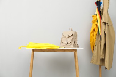 Photo of Yellow umbrella and backpack on white table near rack with coats against light grey background