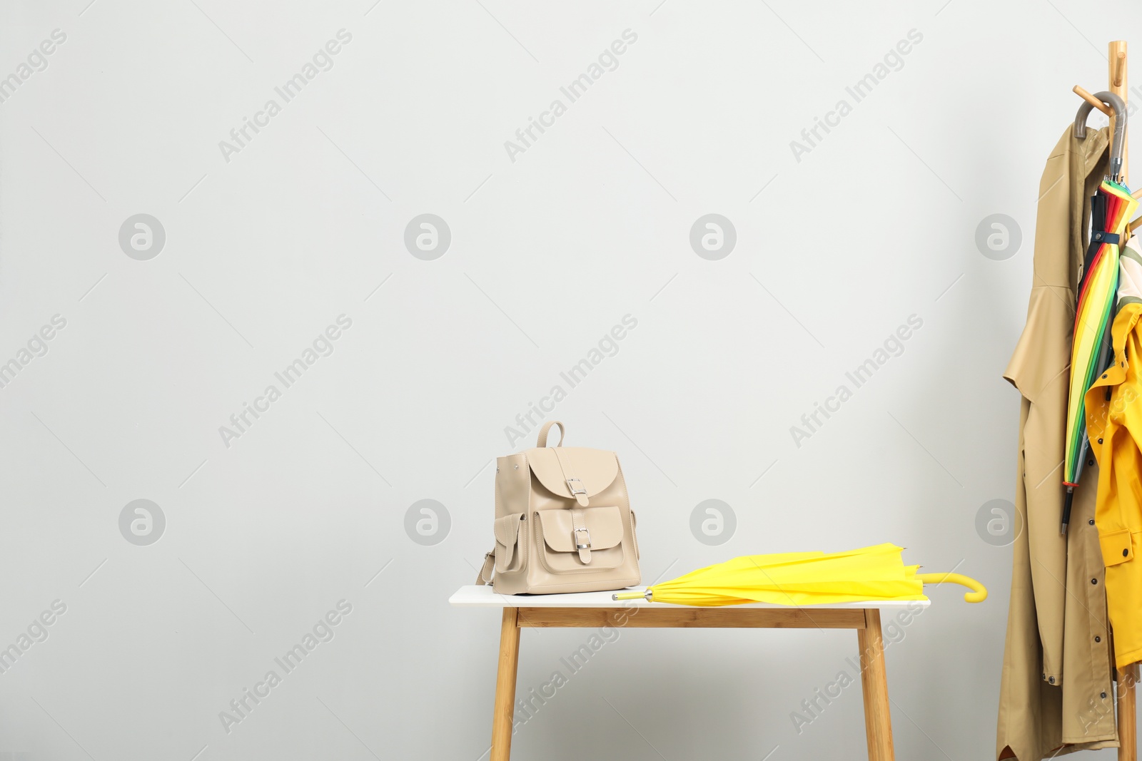 Photo of Yellow umbrella and backpack on white table near rack with coats against light grey background, space for text