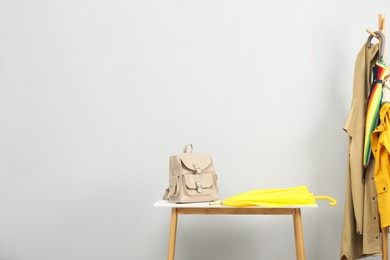 Yellow umbrella and backpack on white table near rack with coats against light grey background, space for text