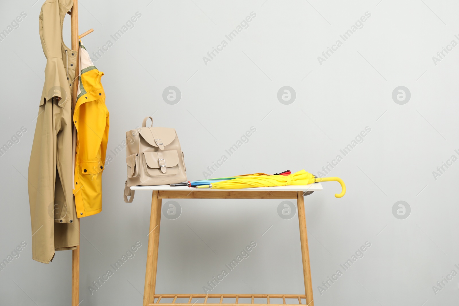 Photo of Colorful umbrellas and backpack on storage bench near rack with coats against light grey background