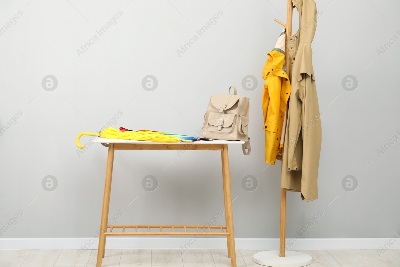 Photo of Colorful umbrellas and backpack on storage bench near rack with coats against light grey background