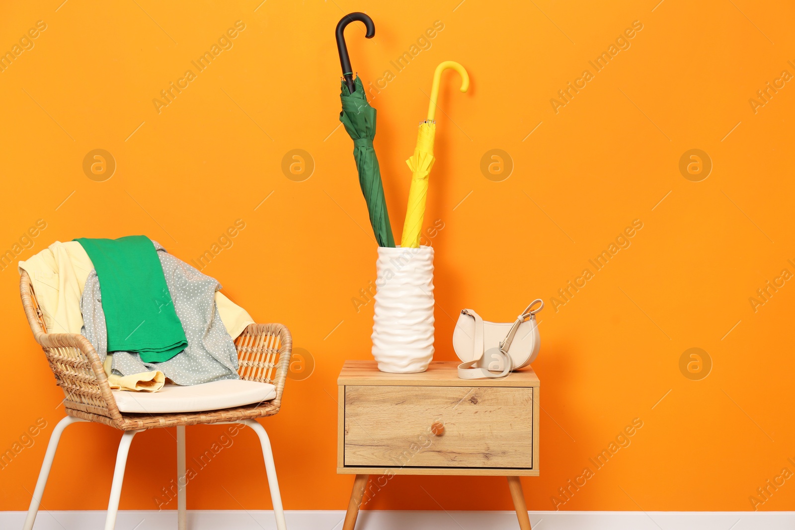 Photo of Colorful umbrellas in holder, bag and clothes near orange wall indoors