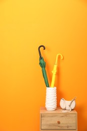 Colorful umbrellas in holder and bag on wooden side table near orange wall indoors