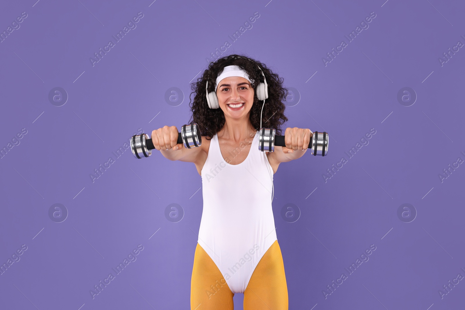 Photo of Woman in headphones doing aerobic exercise with dumbbells on purple background