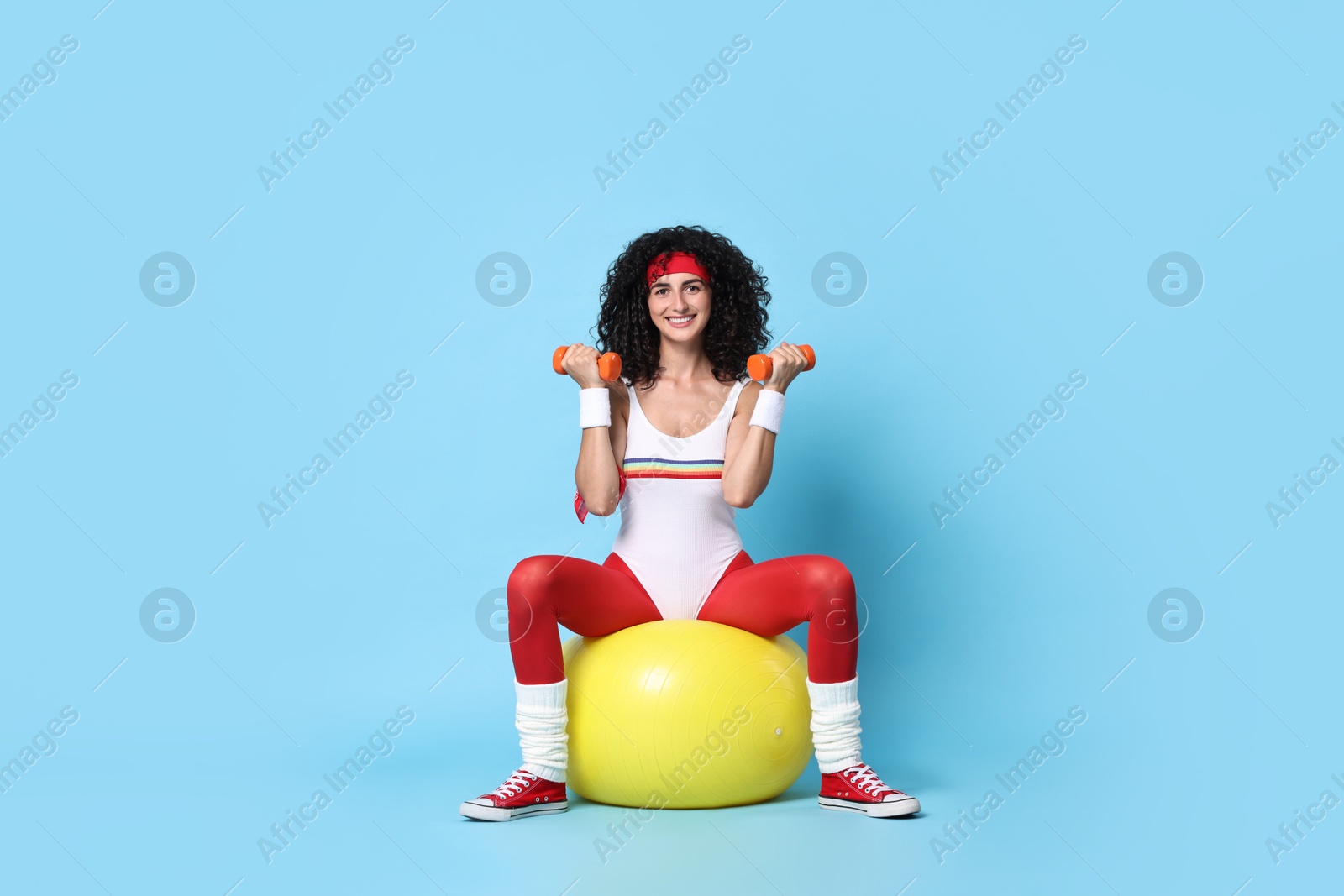 Photo of Woman doing aerobic exercise with dumbbells and fitness ball on light blue background