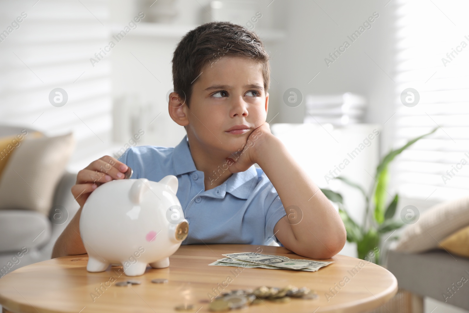 Photo of Pocket money. Cute boy putting coin into piggy bank at wooden table indoors