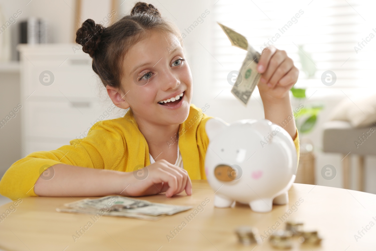 Photo of Pocket money. Cute girl putting dollar banknote into piggy bank at wooden table indoors