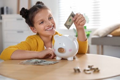 Photo of Pocket money. Cute girl putting dollar banknote into piggy bank at wooden table indoors