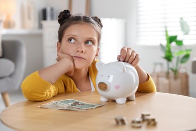 Pocket money. Cute girl putting coin into piggy bank at wooden table indoors