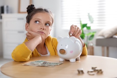 Photo of Pocket money. Cute girl putting coin into piggy bank at wooden table indoors