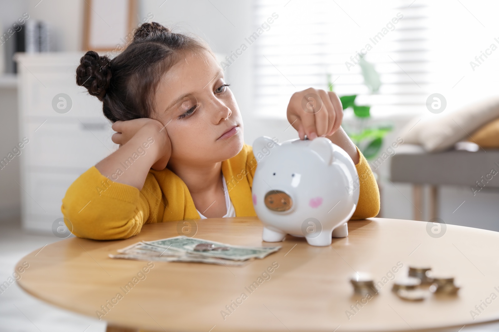 Photo of Pocket money. Cute girl putting coin into piggy bank at wooden table indoors