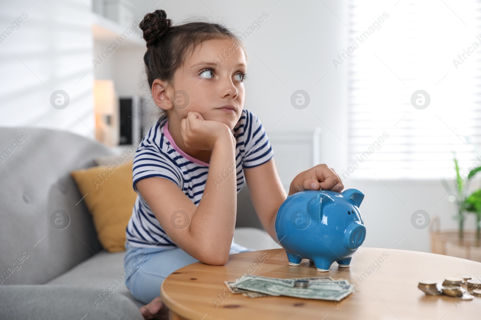 Photo of Pocket money. Cute girl putting coin into piggy bank at wooden table indoors