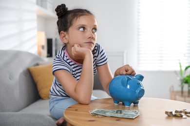 Photo of Pocket money. Cute girl putting coin into piggy bank at wooden table indoors