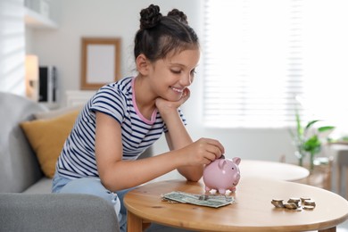 Pocket money. Cute girl putting coin into piggy bank at wooden table indoors