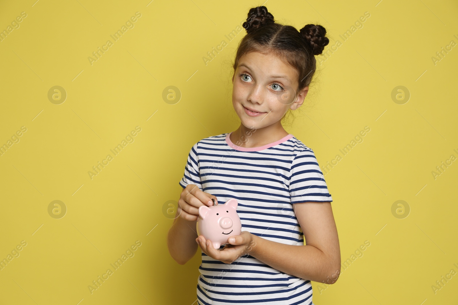 Photo of Pocket money. Cute girl putting coin into piggy bank on yellow background