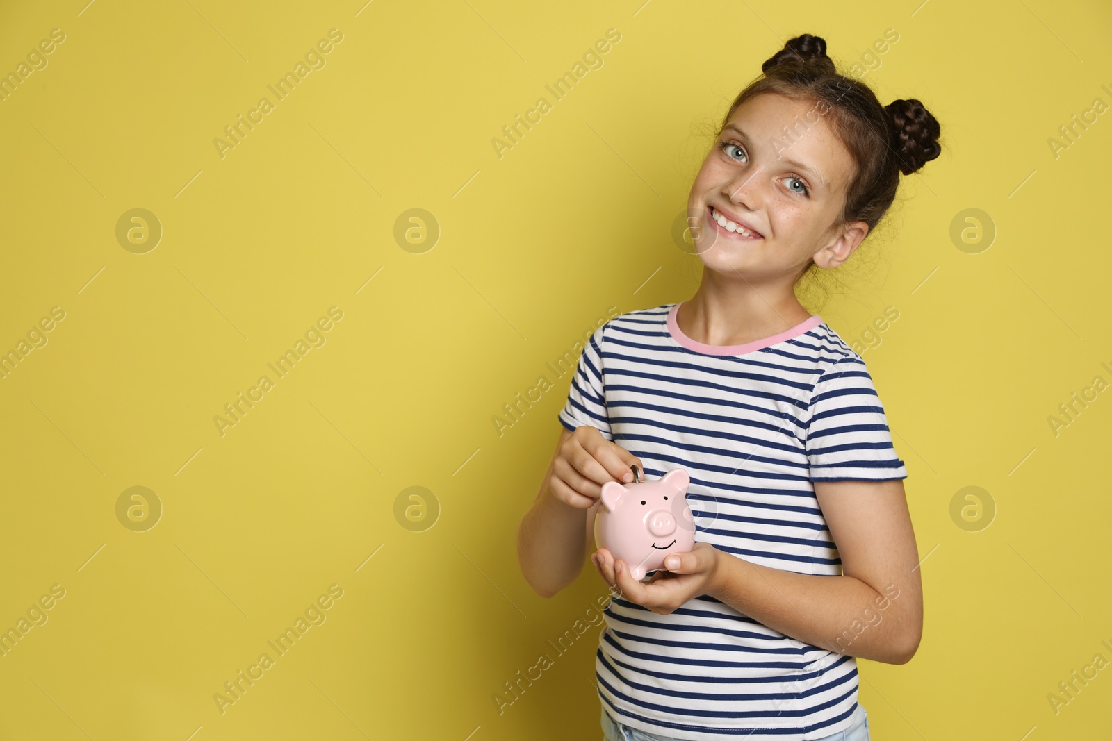 Photo of Pocket money. Cute girl putting coin into piggy bank on yellow background, space for text