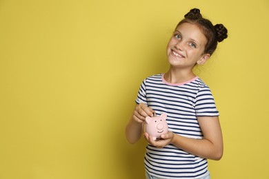 Photo of Pocket money. Cute girl putting coin into piggy bank on yellow background, space for text