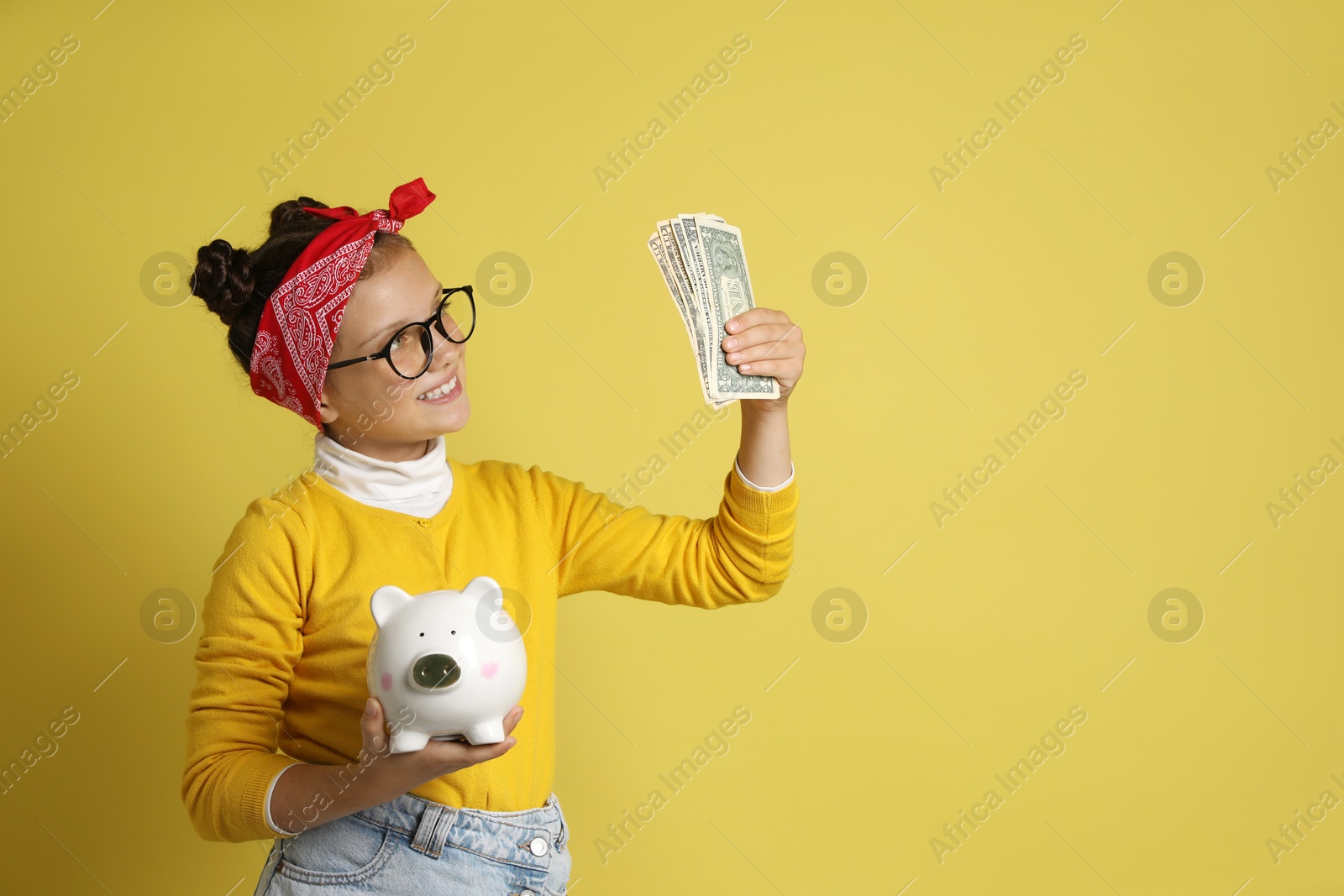 Photo of Pocket money. Cute girl with piggy bank and dollar banknotes on yellow background, space for text