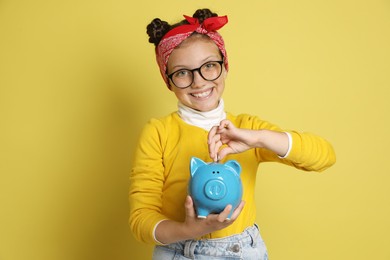 Photo of Pocket money. Cute girl putting coin into piggy bank on yellow background