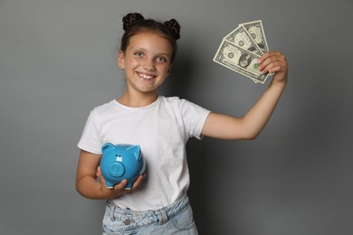 Photo of Pocket money. Cute girl with piggy bank and dollar banknotes on grey background