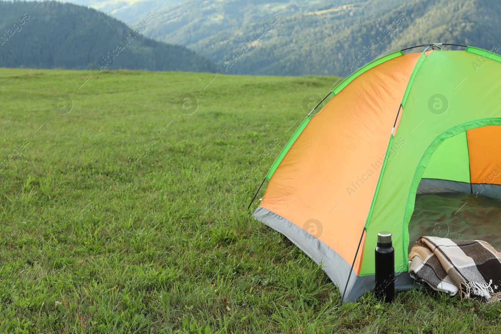 Photo of Tent, thermos and blanket on green grass in mountains, space for text