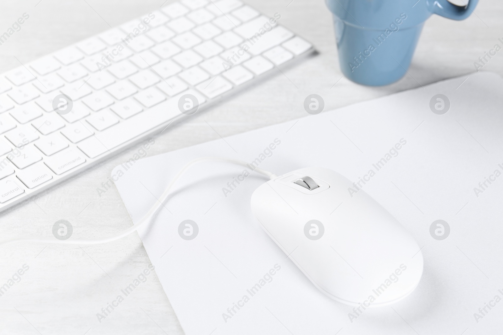 Photo of Computer mouse with mousepad, keyboard and cup on light wooden table, closeup