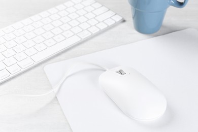 Photo of Computer mouse with mousepad, keyboard and cup on light wooden table, closeup