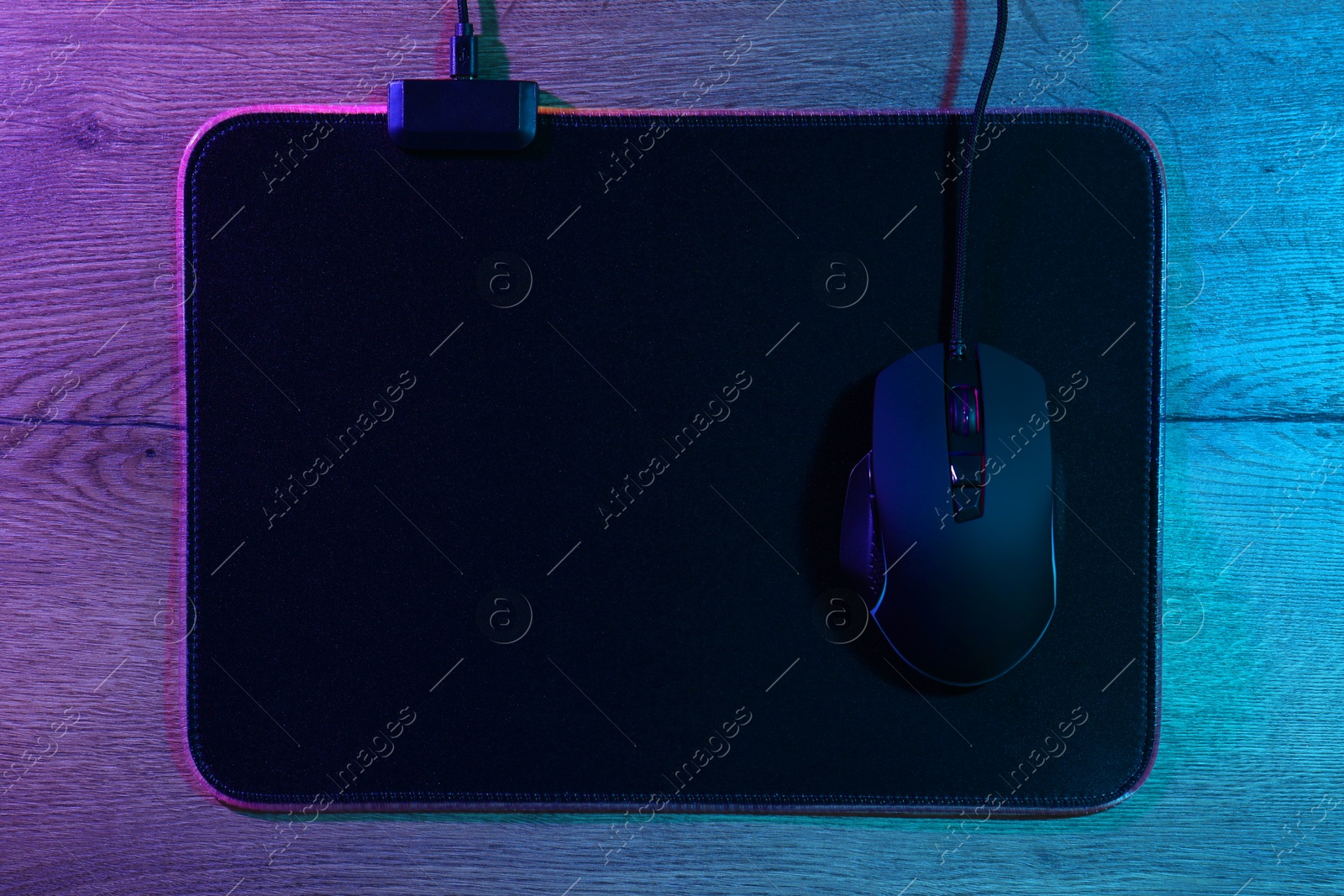 Photo of Black computer mouse and mousepad on wooden table in neon lights, top view