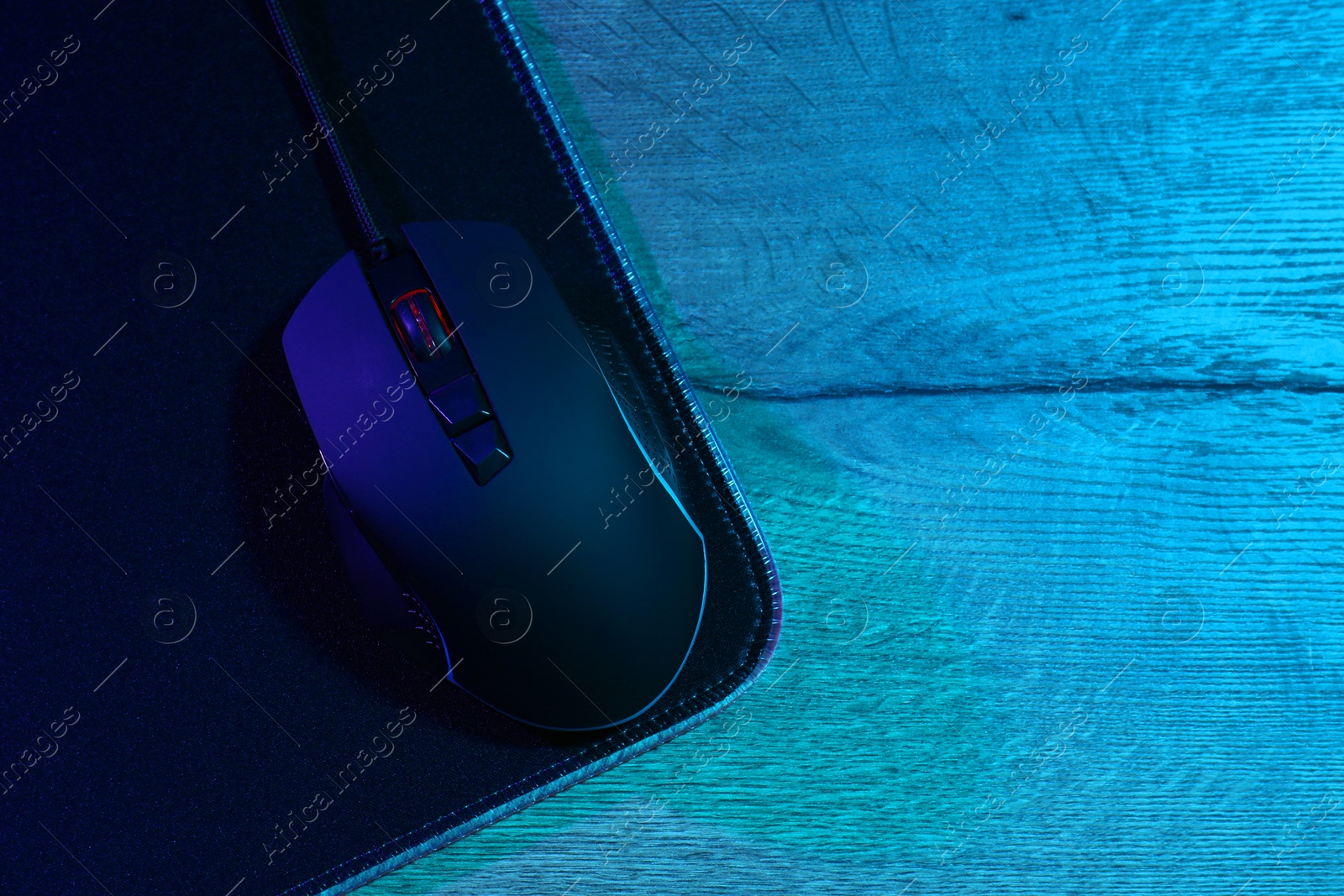 Photo of Black computer mouse and mousepad on wooden table in neon lights, top view. Space for text