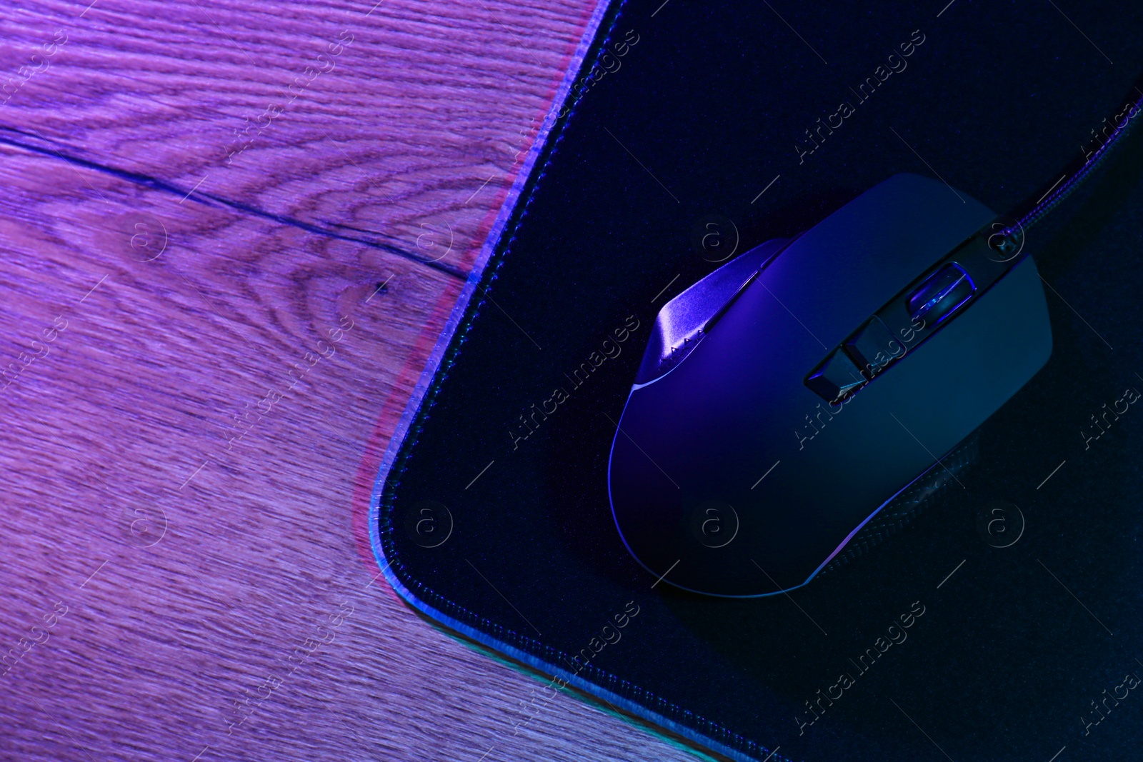 Photo of Black computer mouse and mousepad on wooden table in neon lights, top view. Space for text