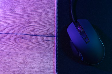 Photo of Black computer mouse and mousepad on wooden table in neon lights, top view. Space for text