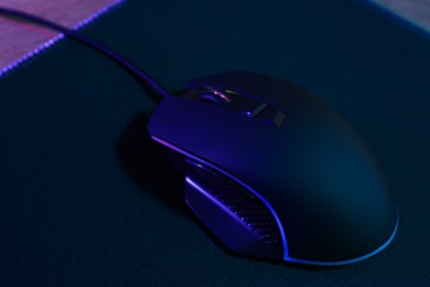 Photo of Black computer mouse and mousepad on wooden table in neon lights, closeup