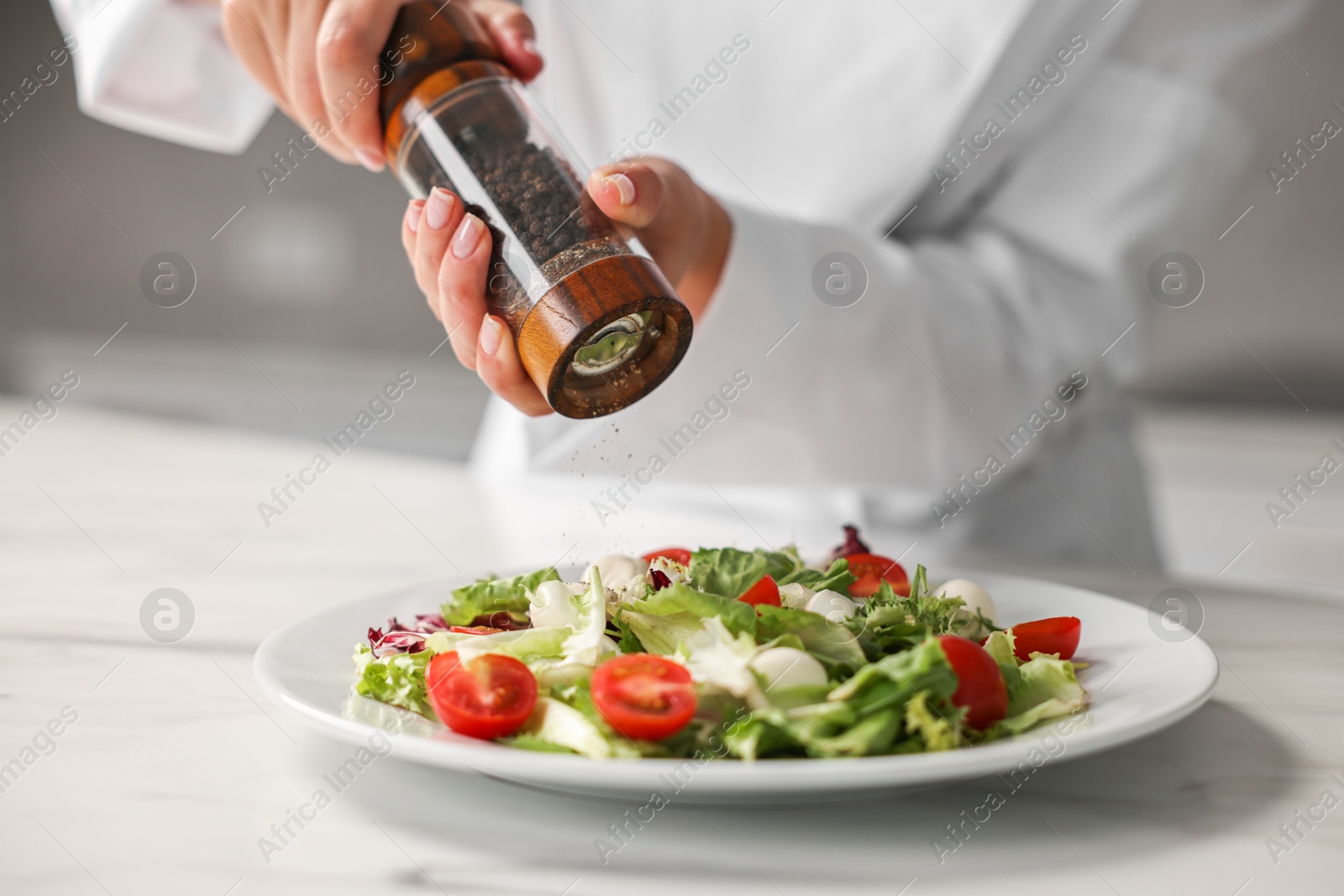 Photo of Professional chef adding pepper to delicious salad at white marble table in kitchen, closeup