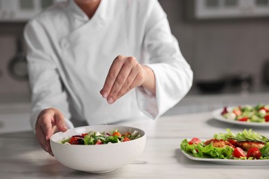 Photo of Professional chef adding salt to delicious salad at white marble table in kitchen, closeup