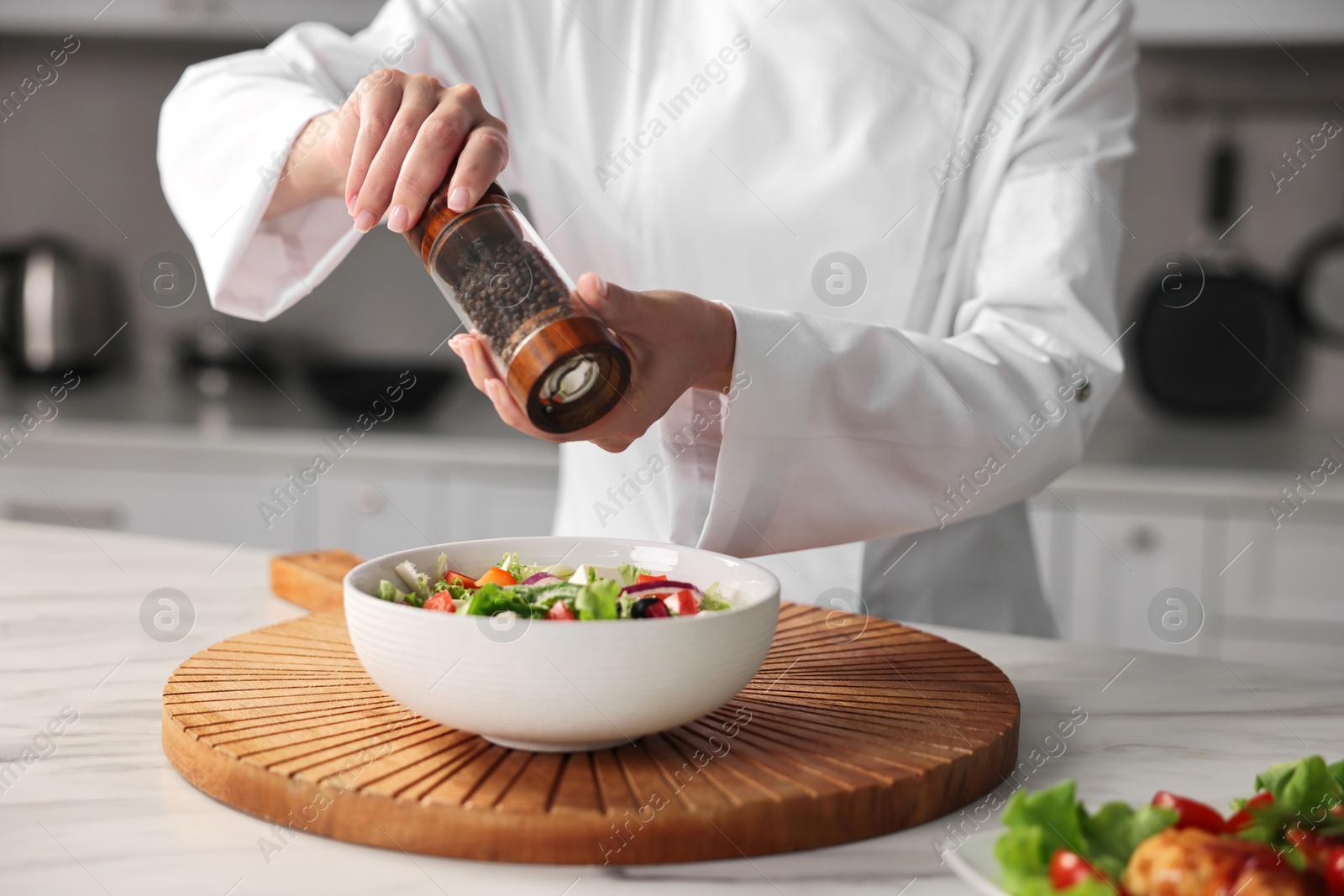 Photo of Professional chef adding pepper to delicious salad at white marble table in kitchen, closeup