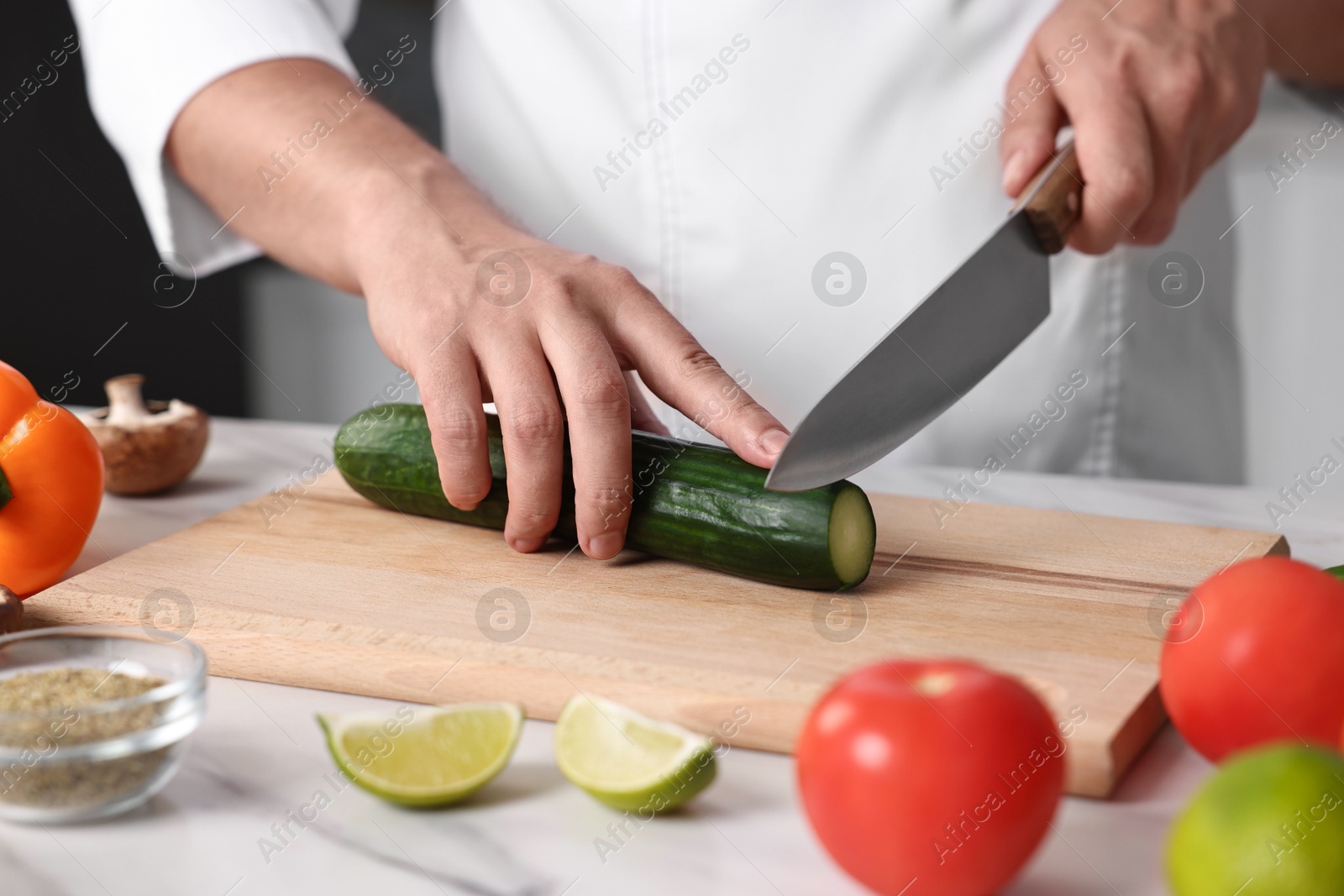 Photo of Professional chef cutting cucumber at white marble table in kitchen, closeup