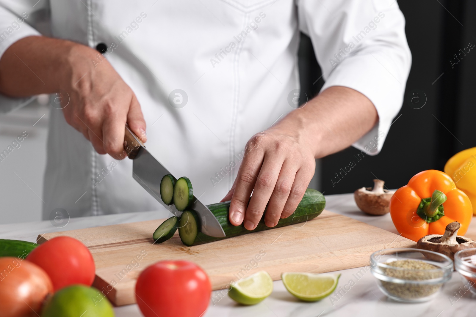 Photo of Professional chef cutting cucumber at white marble table in kitchen, closeup