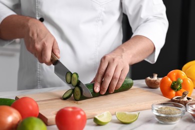 Professional chef cutting cucumber at white marble table in kitchen, closeup