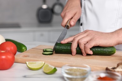 Professional chef cutting cucumber at white marble table in kitchen, closeup