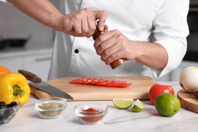 Photo of Professional chef adding salt to tomato at white marble table in kitchen, closeup