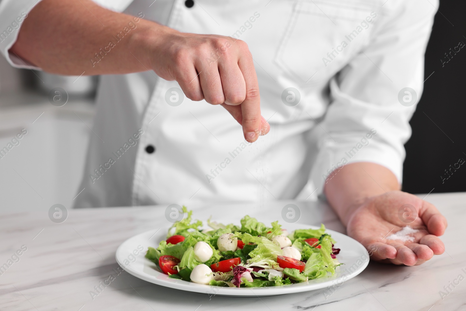 Photo of Professional chef adding salt to delicious salad at white marble table in kitchen, closeup