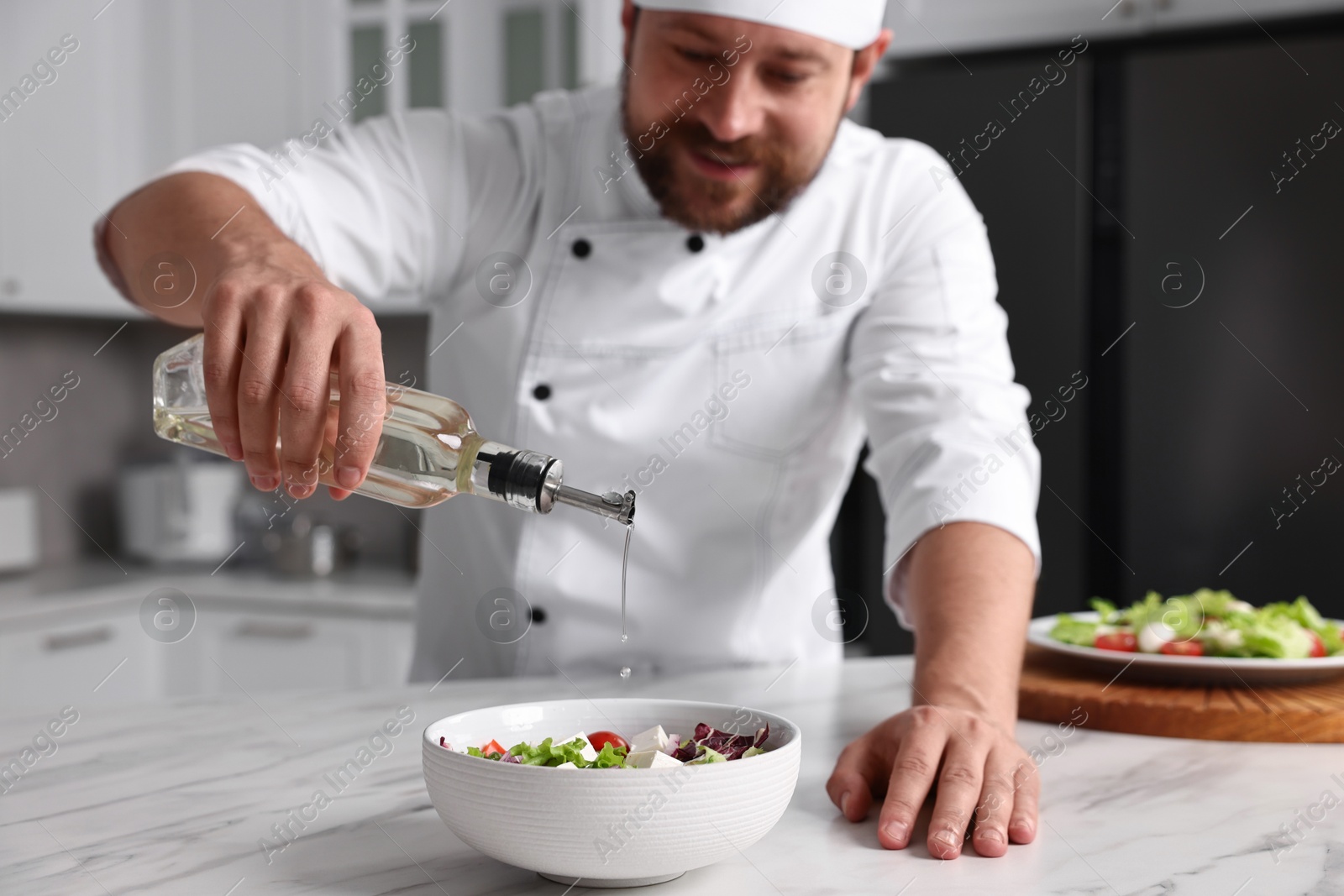 Photo of Professional chef pouring oil onto delicious salad at white marble table in kitchen, selective focus